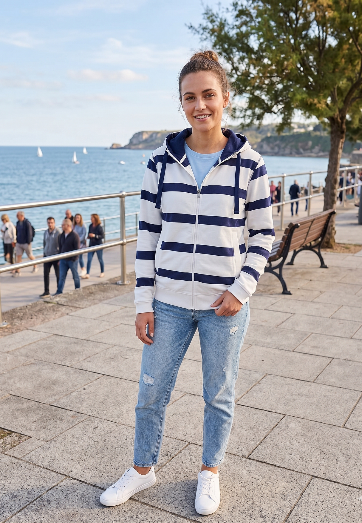 Person wearing a striped hoodie and jeans standing by a waterfront with people and trees in the background.