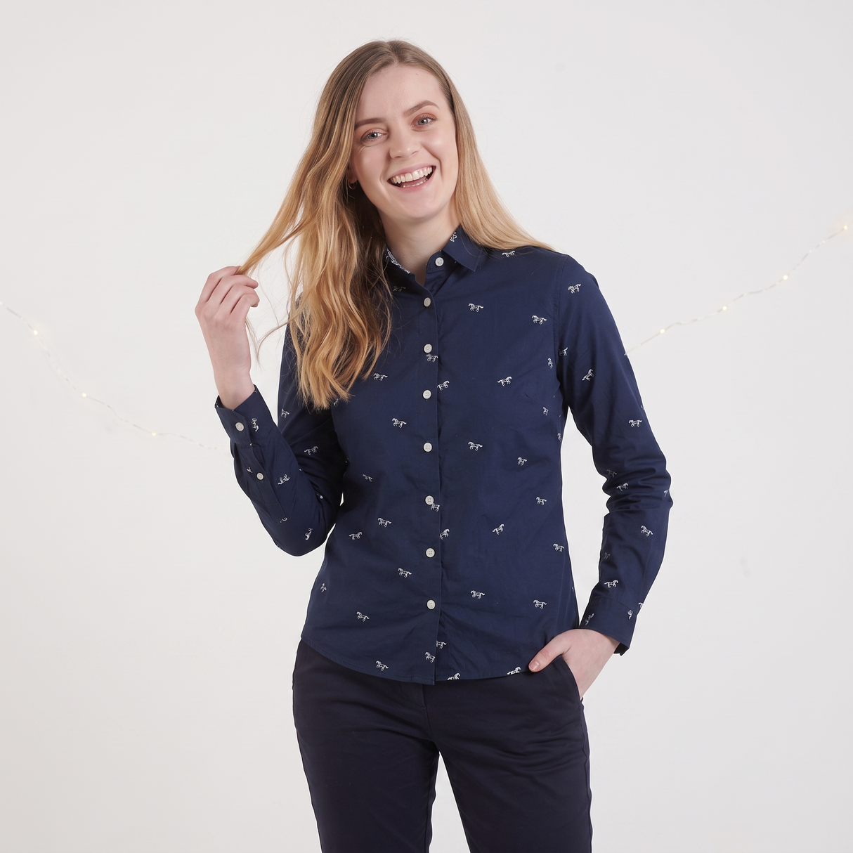 A woman modeling a navy blue long-sleeve button-up shirt with a small white allover pattern, paired with dark trousers, against a light gray studio background.