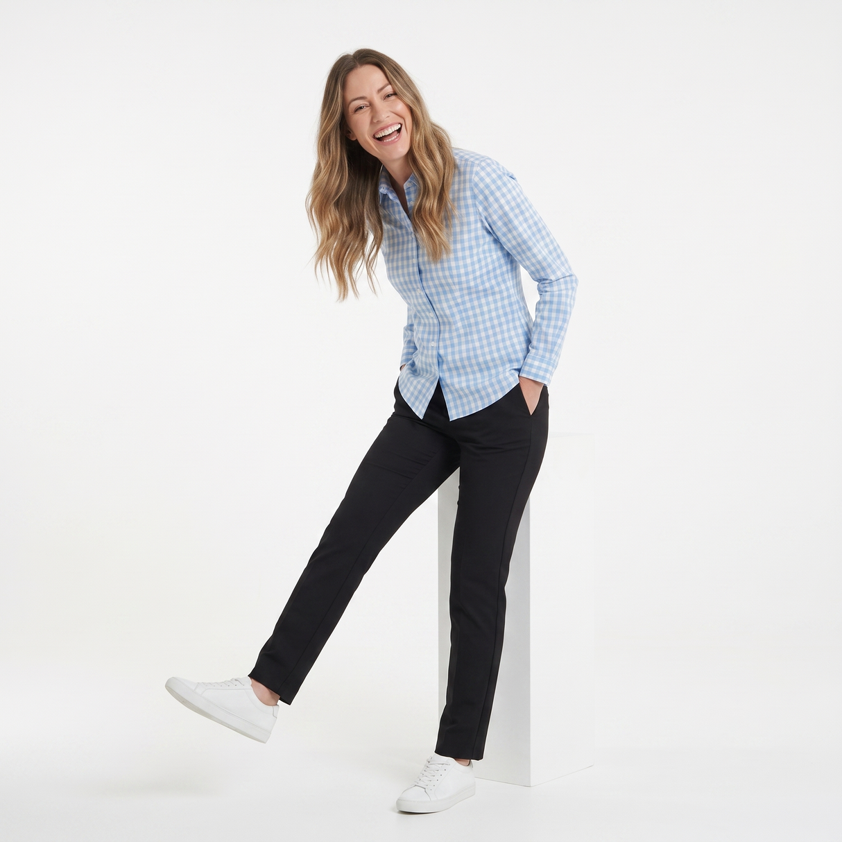 A woman modeling a light blue and white gingham long-sleeve button-down shirt, paired with black trousers and white sneakers, posing on a white studio pedestal.
