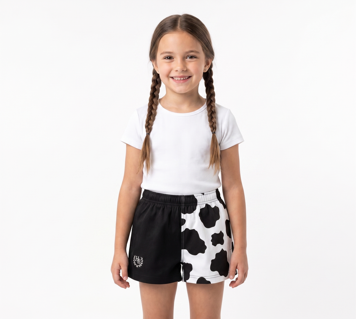 A young girl with braided hair stands against a white studio background wearing a white t-shirt and black-and-white cow-print shorts.
