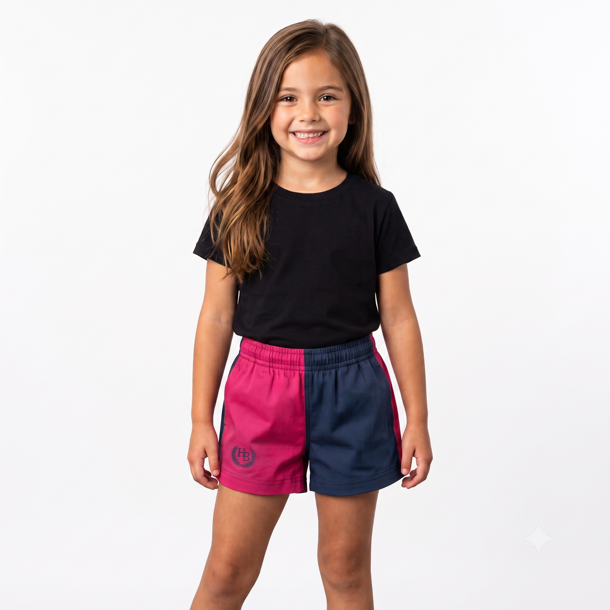 A young girl with long brown hair stands against a plain white studio background, wearing a black short-sleeve t-shirt and athletic shorts with a split design of bright pink and navy blue. A circular logo is visible on the pink leg.
