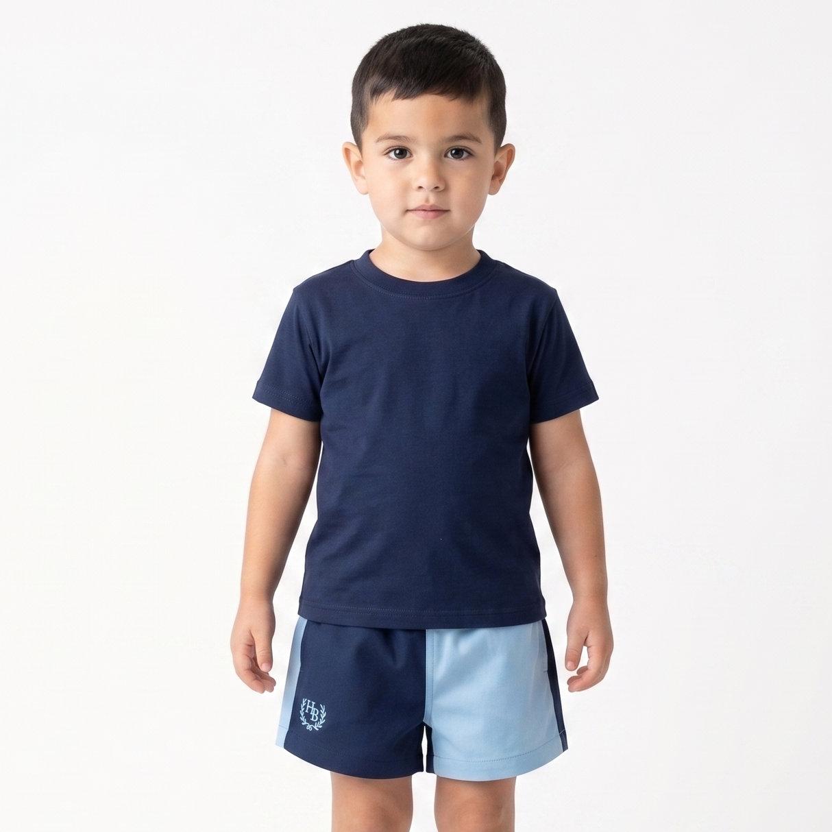 A young boy stands barefoot against a plain white studio background, wearing a navy blue short-sleeve t-shirt and matching navy shorts with a light blue side panel and a small white crest logo near the left hem.