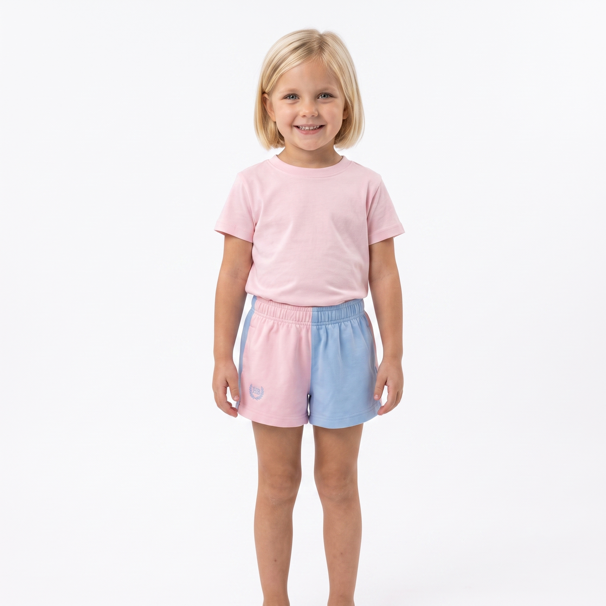 A young girl with blonde hair stands against a white studio background wearing a light pink t-shirt and pastel split-color shorts in pink and light blue.