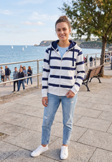 Person wearing a striped hoodie and jeans standing by a waterfront with people and trees in the background.
