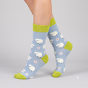 A pair of crew socks worn on a person's feet, featuring a light blue background with white sheep and daisy patterns, bright lime green cuffs, heels, and toes, and small black eyes on the sheep.