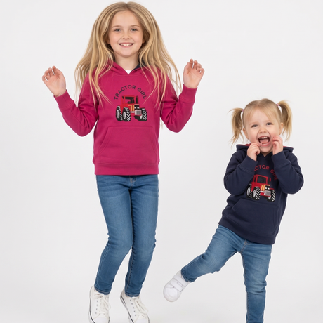 Two young girls jumping against a white studio background, wearing matching Tractor Girl brand hoodies. The older girl wears a bright pink hoodie with a red tractor graphic, and the younger girl wears a navy hoodie with a red tractor graphic.