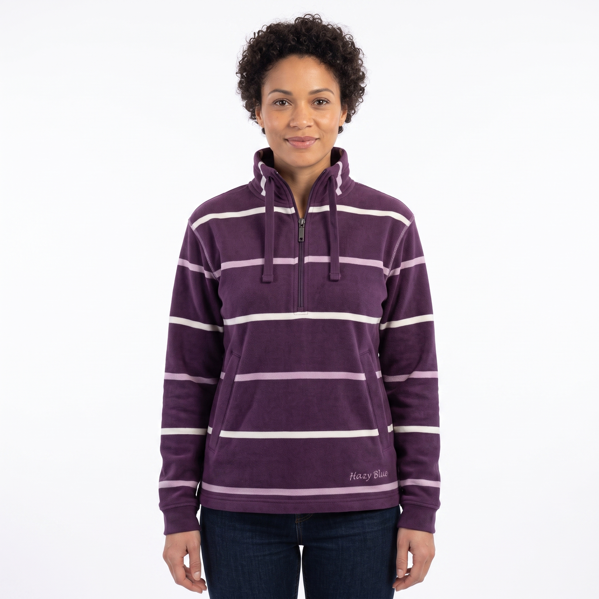 A woman modeling a purple half-zip fleece pullover with white horizontal stripes, featuring a drawstring neck and a small 'Hazy Blue' brand logo near the bottom right hem, set against a plain white background.