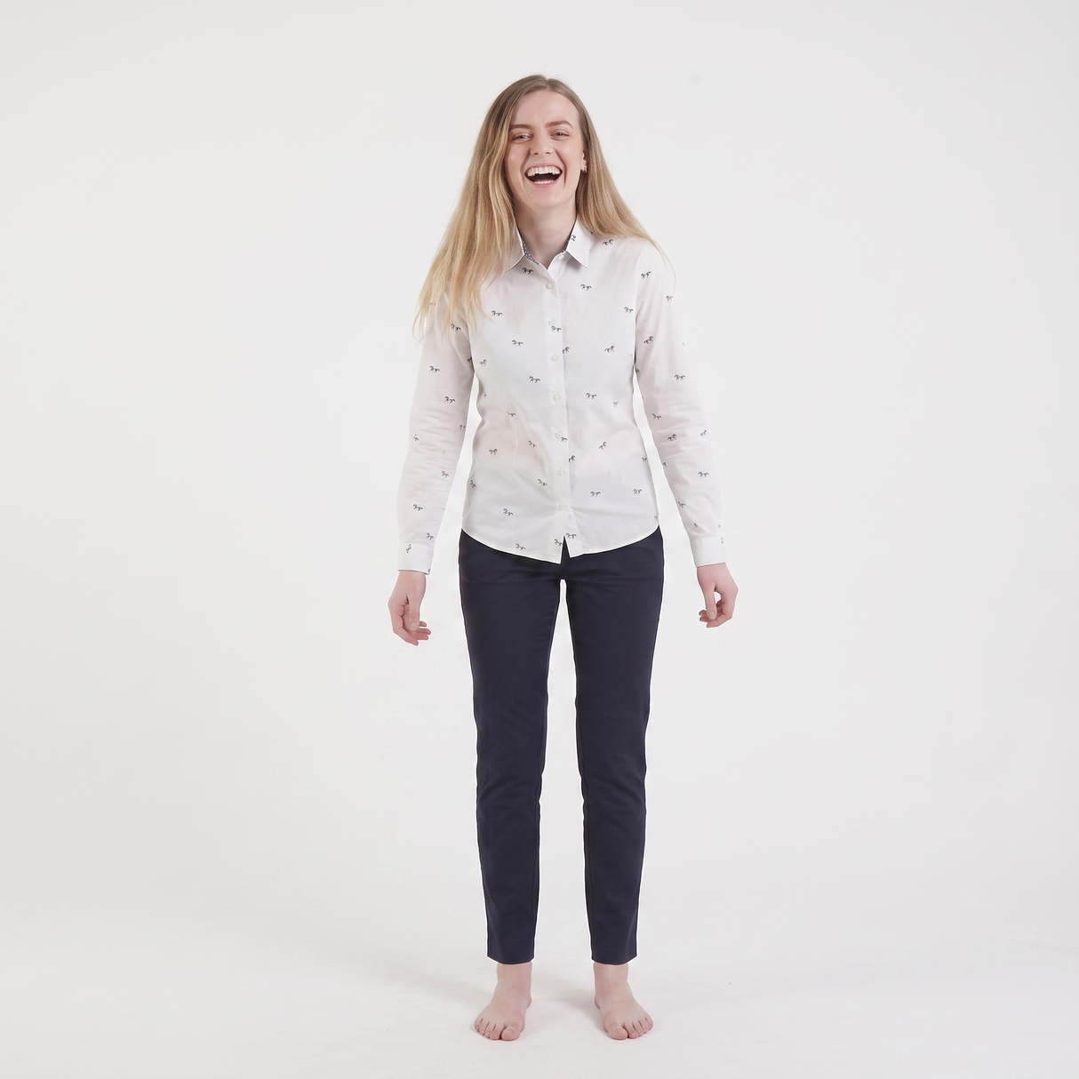 A young woman with blonde hair laughing, wearing a white long-sleeve button-down shirt with a small blue and pink pattern, paired with dark navy pants, standing against a solid white studio background.