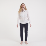 A young woman with blonde hair laughing, wearing a white long-sleeve button-down shirt with a small blue and pink pattern, paired with dark navy pants, standing against a solid white studio background.