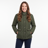 A smiling woman with long brown hair models an olive green diamond-quilted jacket with a high collar and front buttons, paired with navy blue pants, against a plain white studio background.
