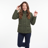 A woman smiling and posing in a quilted olive green jacket with dark navy pants, set against a plain white studio background.