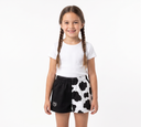 A young girl with braided hair stands against a white studio background wearing a white t-shirt and black-and-white cow-print shorts.