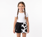 A young girl with braided hair stands against a white studio background wearing a white t-shirt and black-and-white cow-print shorts.