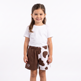 A young girl stands against a plain white studio background wearing a white t-shirt and brown shorts with white cow-print spots. A small circular logo is visible on the lower left leg of the shorts.