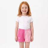 A young girl with red hair stands against a plain white background wearing a white t-shirt and pink shorts with a split-color design, one half dark pink and one half light pink.