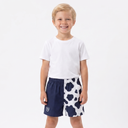 A young boy stands barefoot against a plain white studio background, wearing a white short-sleeve T-shirt and shorts with a split design: one half is solid navy blue and the other half is white with navy cow-print spots.