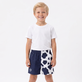 A young boy stands barefoot against a plain white studio background, wearing a white short-sleeve T-shirt and shorts with a split design: one half is solid navy blue and the other half is white with navy cow-print spots.