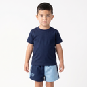 A young boy stands barefoot against a plain white studio background, wearing a navy blue short-sleeve t-shirt and matching navy shorts with a light blue side panel and a small white crest logo near the left hem.
