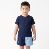 A young boy stands barefoot against a plain white studio background, wearing a navy blue short-sleeve t-shirt and matching navy shorts with a light blue side panel and a small white crest logo near the left hem.