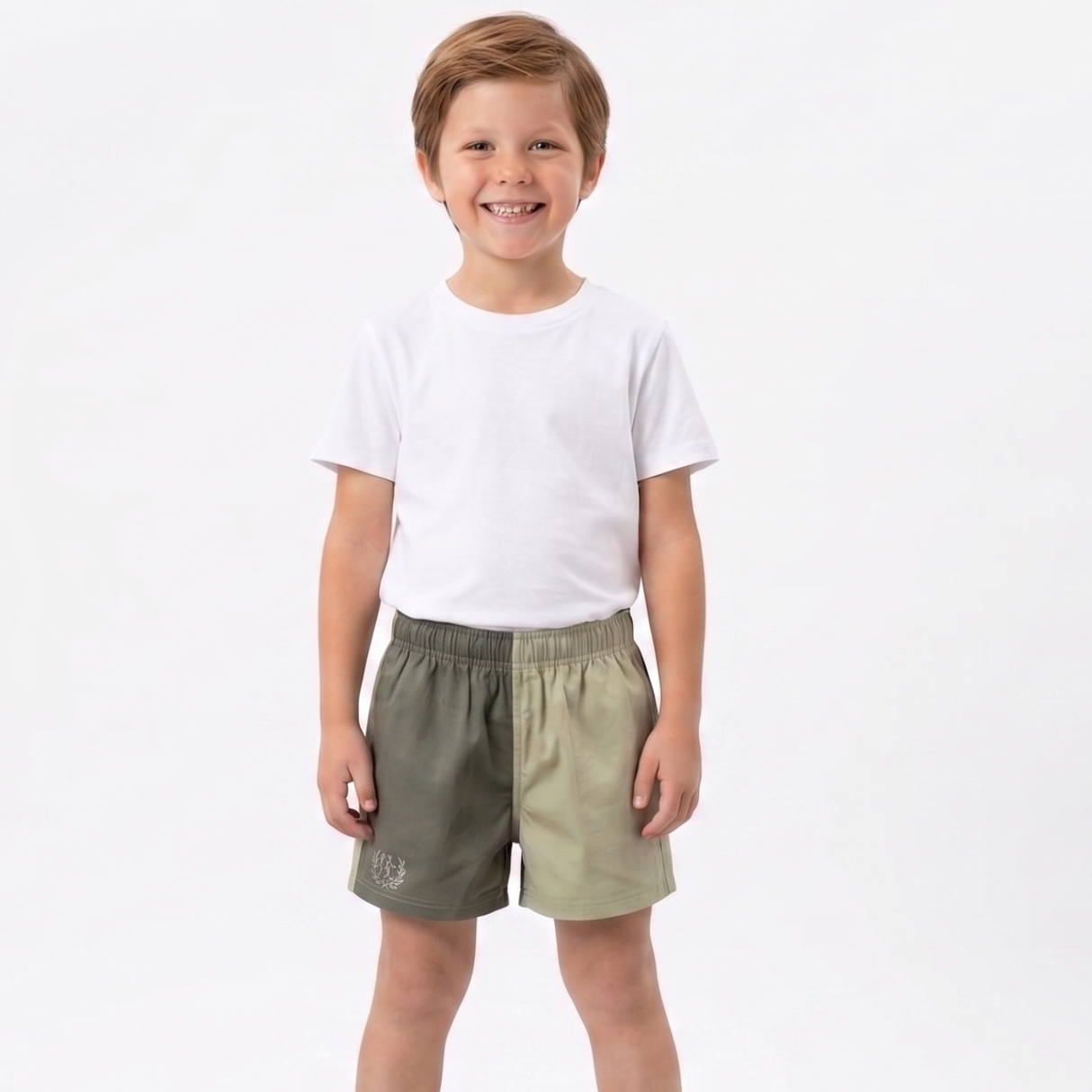 A young boy standing against a white studio background, wearing a white short-sleeve t-shirt and split-color shorts with one leg in olive green and the other in light khaki.