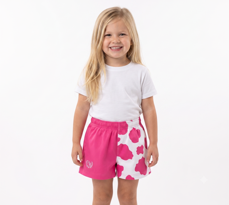 A young girl with long blonde hair stands against a plain white studio background, wearing a white short-sleeve t-shirt and bright pink shorts with a white cloud pattern and a small white crest logo on the left leg.