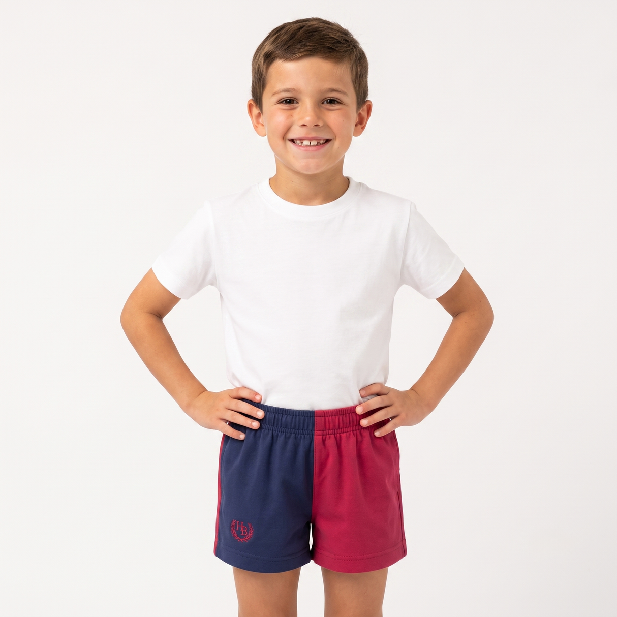 A young boy smiling and posing with hands on hips, wearing a plain white t-shirt and athletic shorts that are split vertically into navy blue on the left and magenta pink on the right, with a small red crest logo on the lower left leg.
