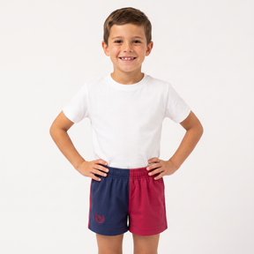 A young boy smiling and posing with hands on hips, wearing a plain white t-shirt and athletic shorts that are split vertically into navy blue on the left and magenta pink on the right, with a small red crest logo on the lower left leg.