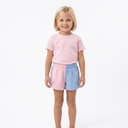 A young girl with blonde hair stands against a white studio background wearing a light pink t-shirt and pastel split-color shorts in pink and light blue.