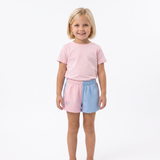 A young girl with blonde hair stands against a white studio background wearing a light pink t-shirt and pastel split-color shorts in pink and light blue.